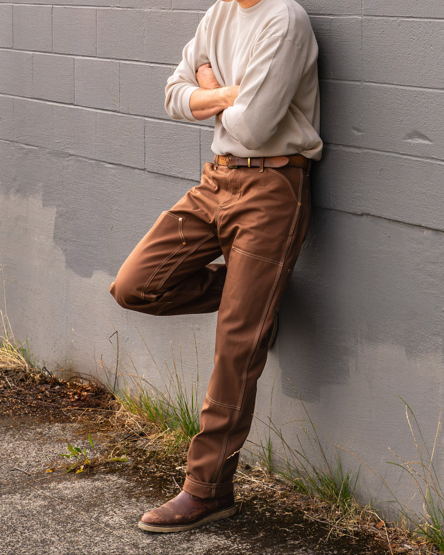 A man wearing a blue baseball cap, cream long-sleeve shirt, brown pants, and brown boots leans against a gray brick wall outdoors.