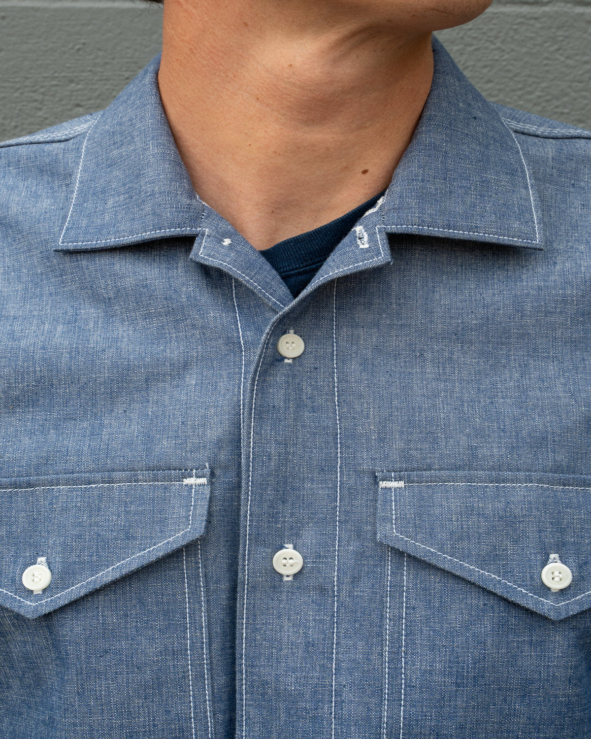 Close-up of a person wearing a blue chambray button-up shirt with two chest pockets and white buttons, layered over a dark navy undershirt.