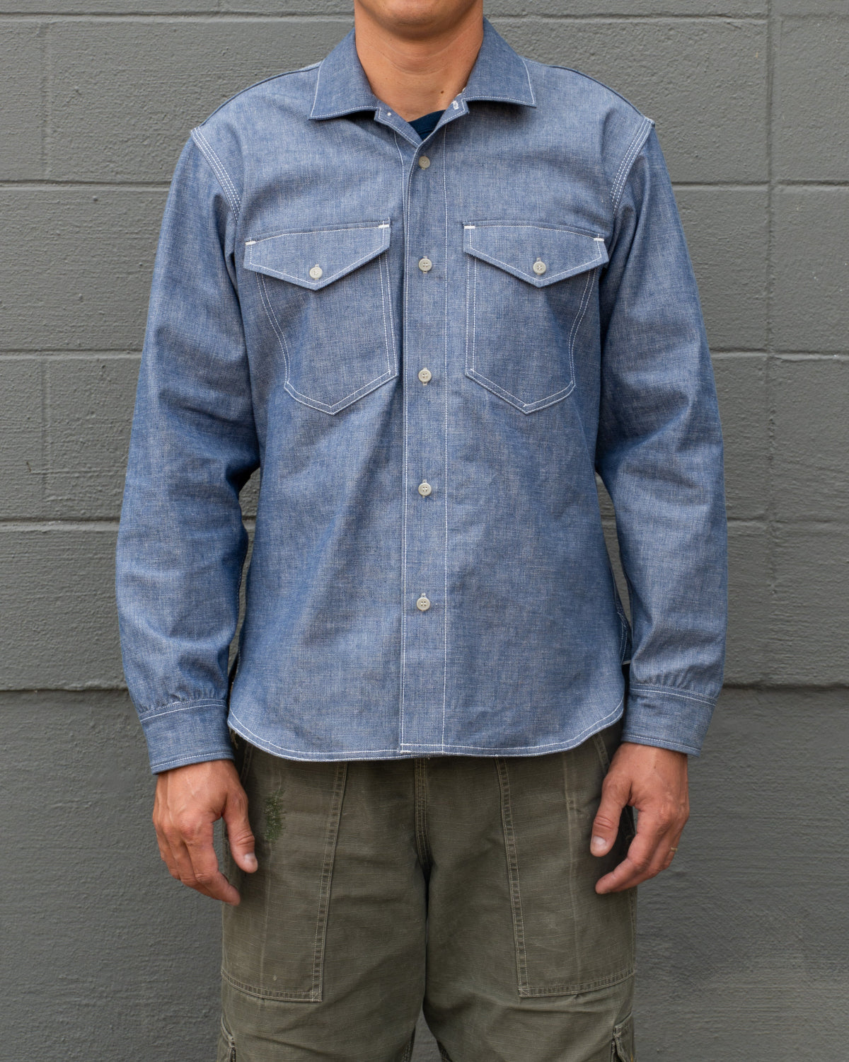 A man wearing a light blue denim long-sleeve button-up shirt with two chest pockets and olive green cargo pants, standing against a gray brick wall.